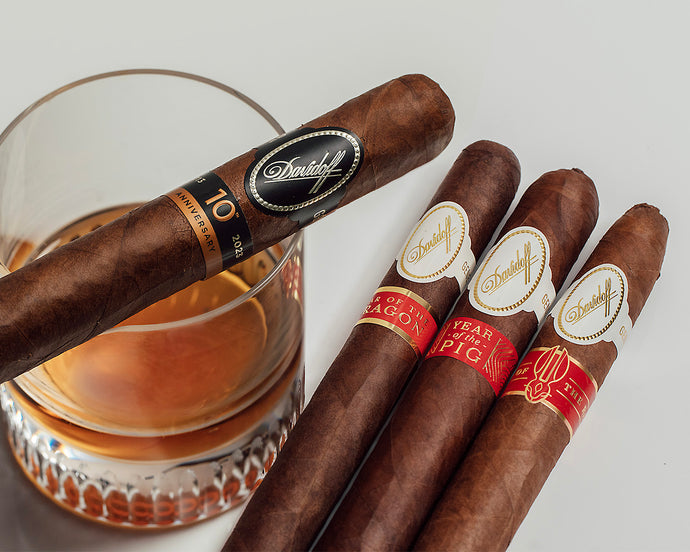 Four Davidoff cigars with decorative bands next to a glass of whiskey on a light gray background
