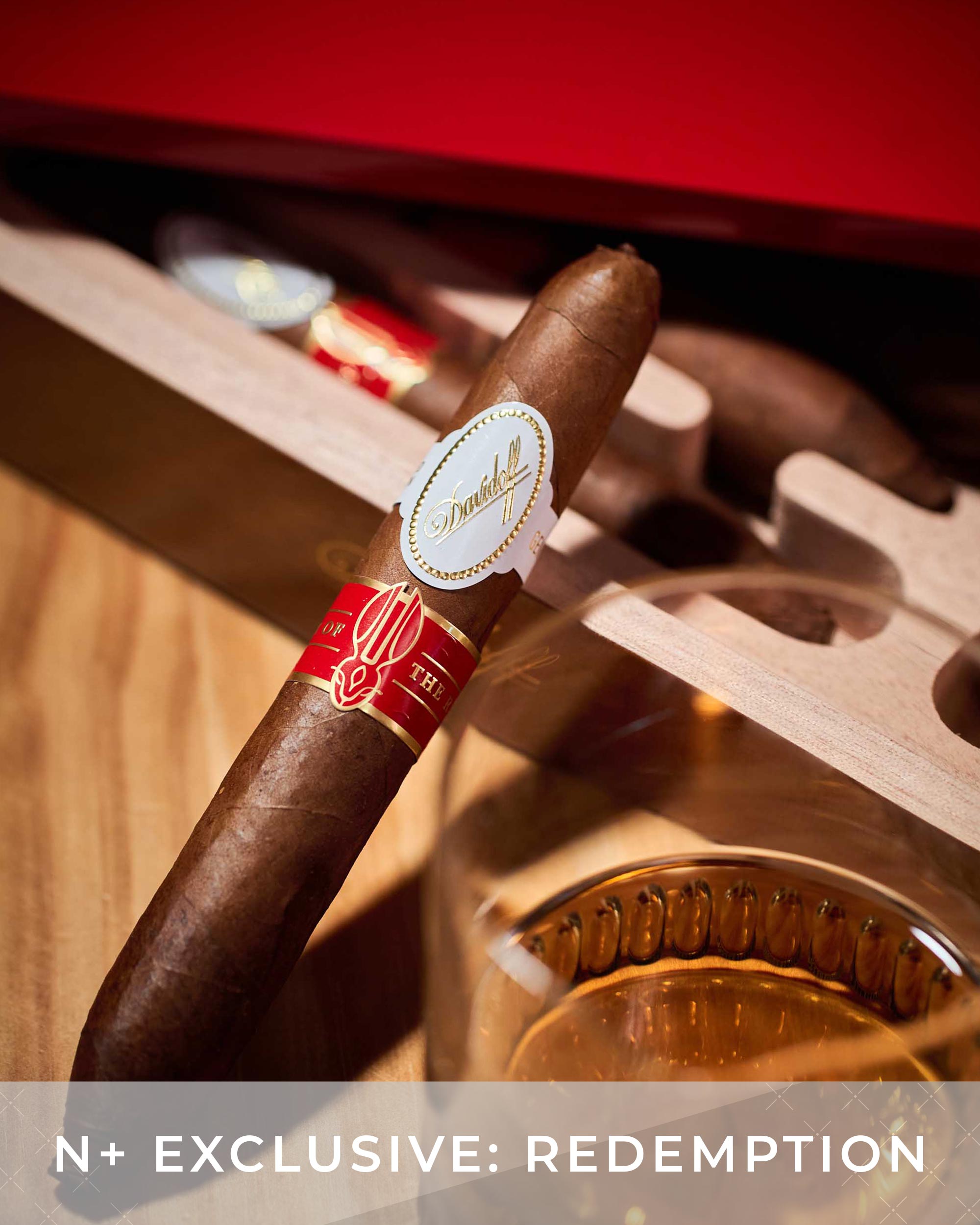 Cigar with a red band and gold label on a wooden stand with a glass of whiskey.