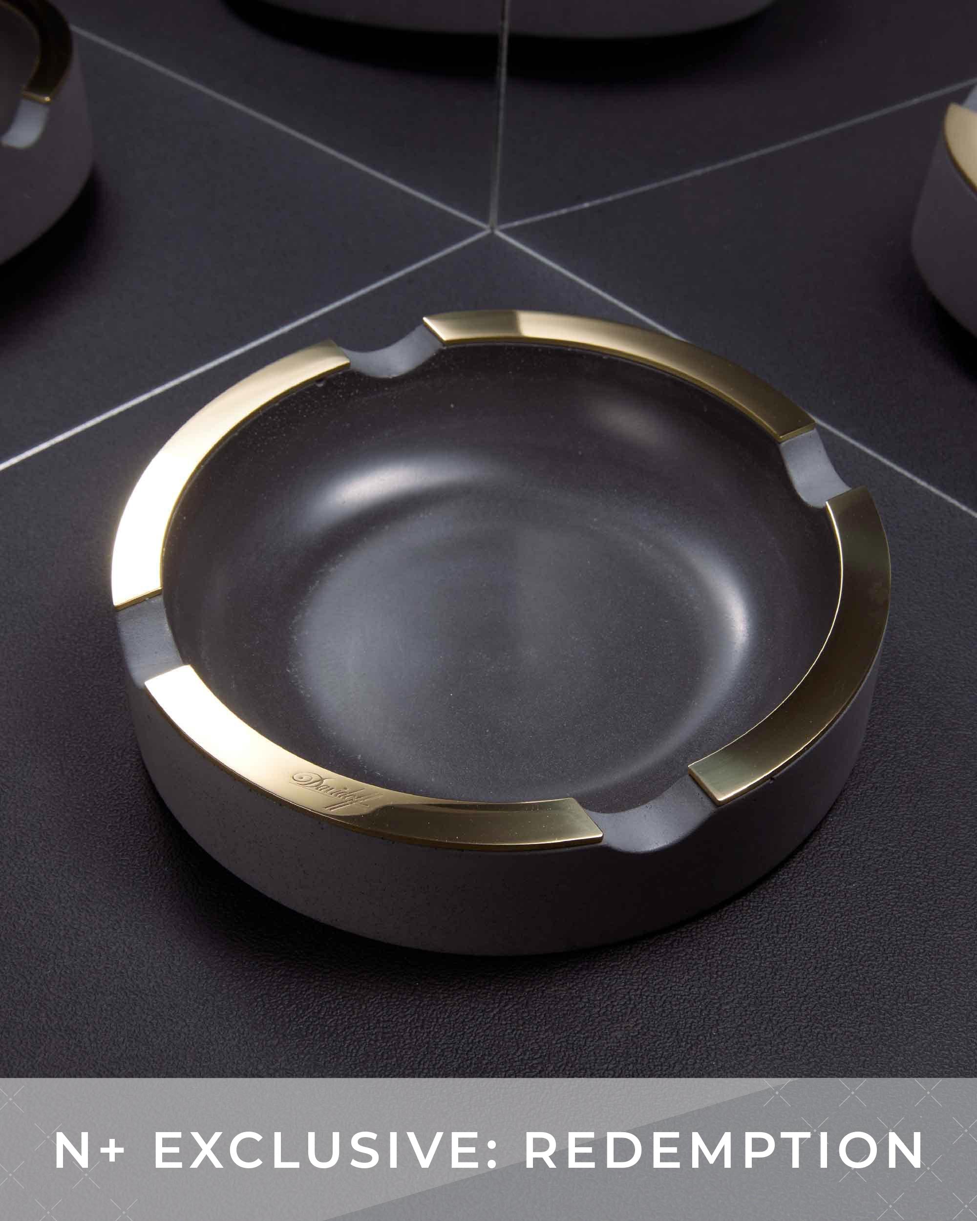 Black ashtray with gold rim on a dark surface, featuring 'N+ Exclusive: Redemption' text.