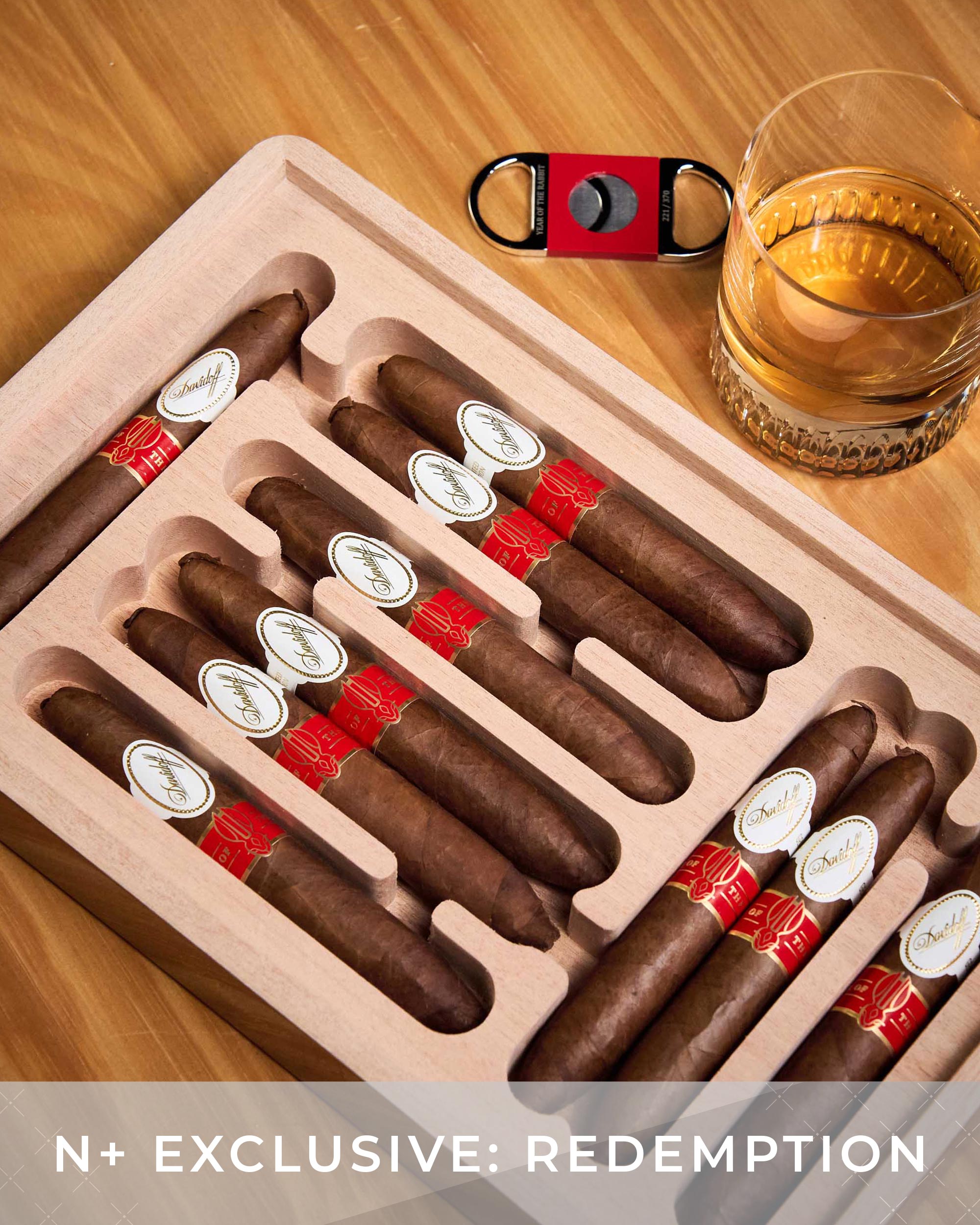 Set of cigars in a wooden humidor with a glass of whiskey and a cutter on a wooden table.