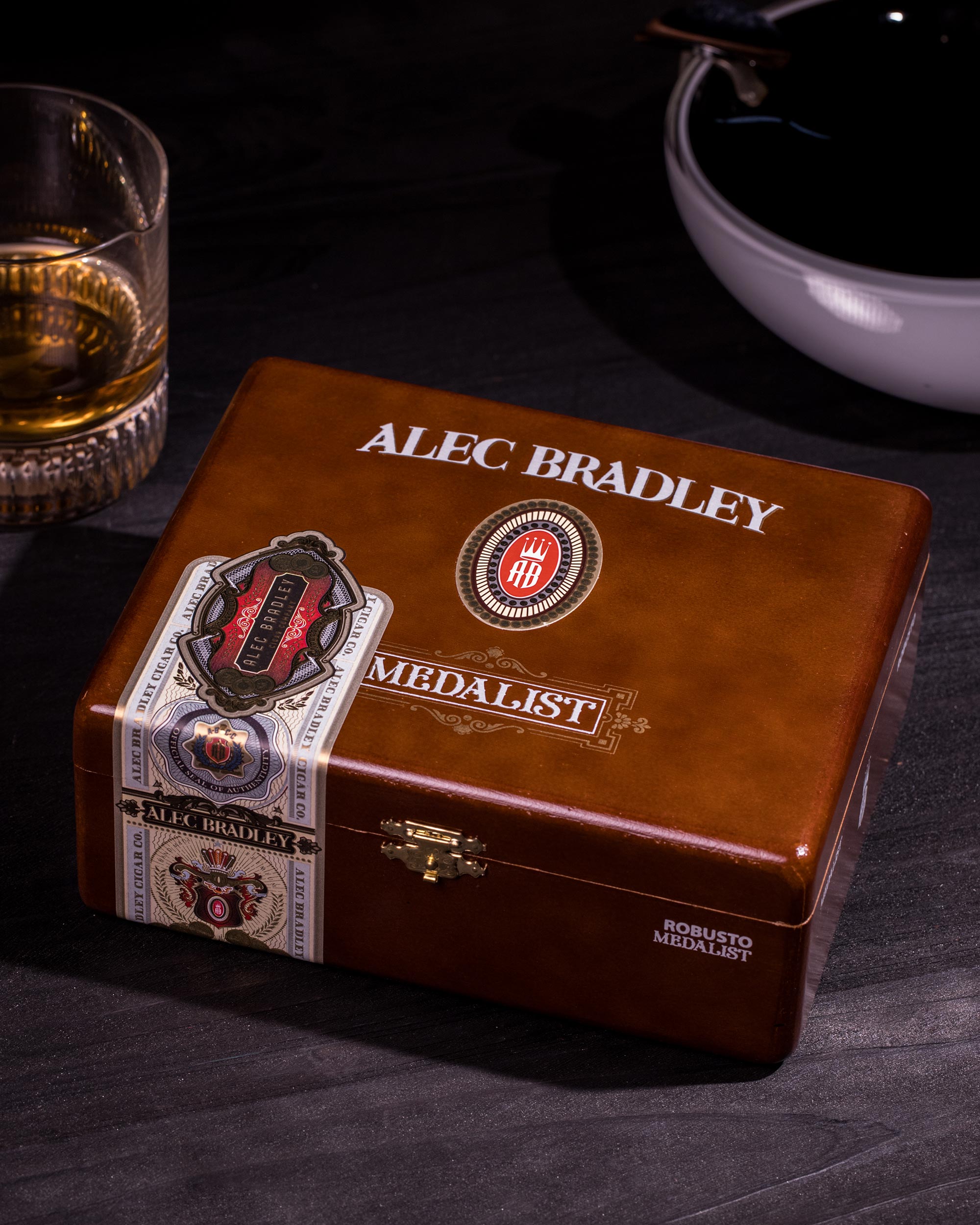 Alec Bradley Medalist cigar box on a dark surface with a glass of whiskey and bowl in the background.