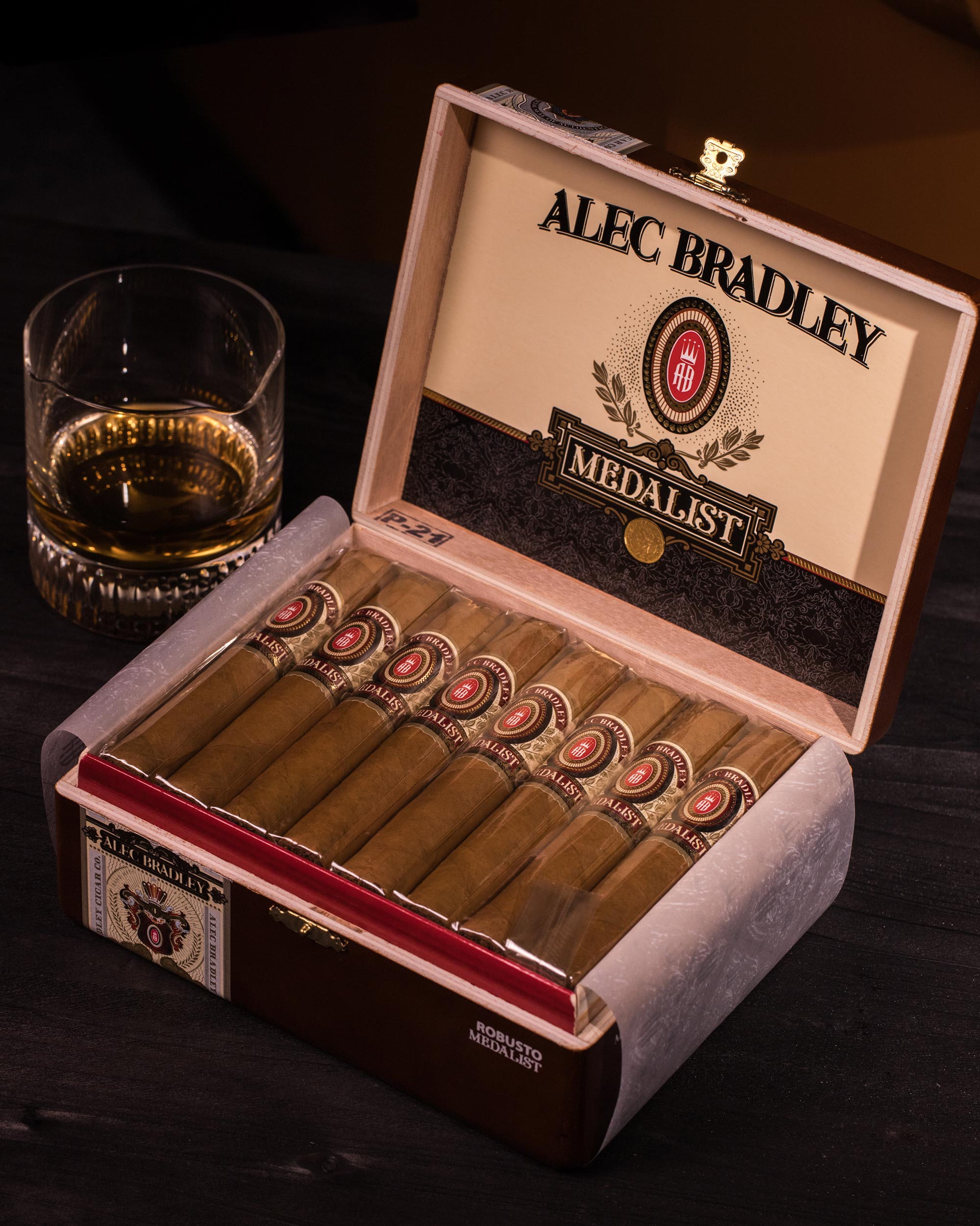 Alec Bradley Medallist cigars in an open humidor with a glass of whiskey on a dark surface.