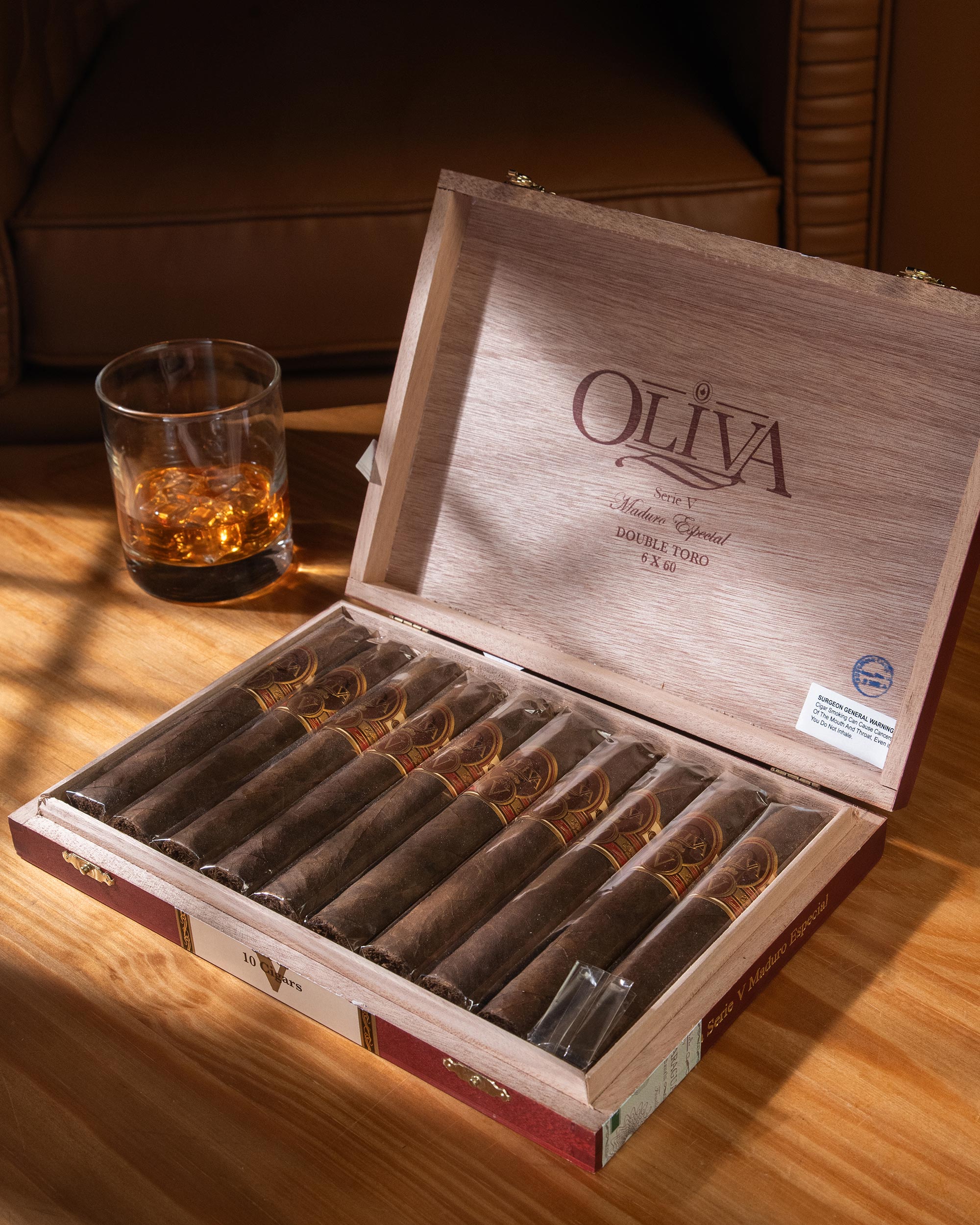 Open wooden box of Oliva cigars with a glass of whiskey on a wooden table.