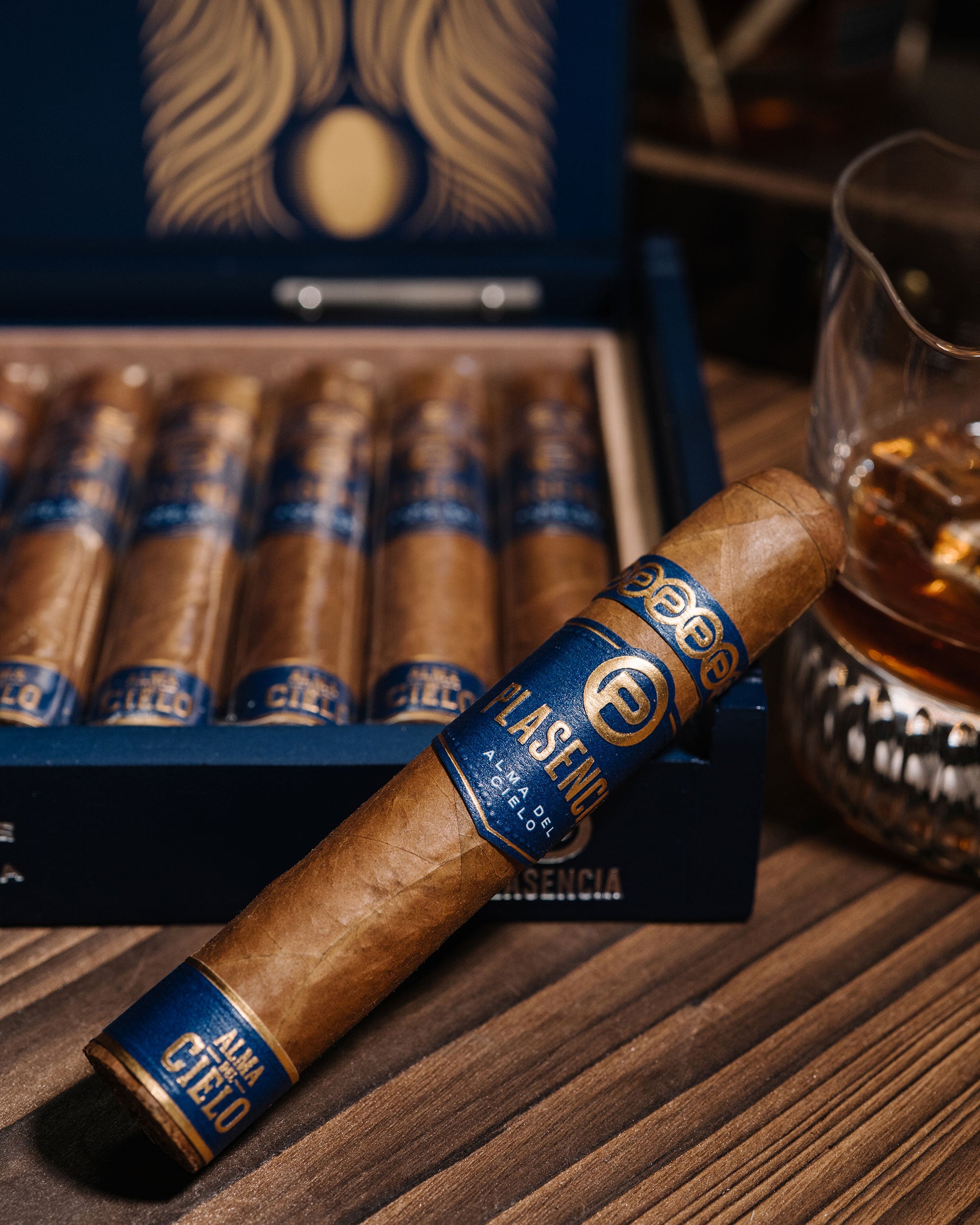 Cigar with blue band in front of a box of cigars and a glass of whiskey.