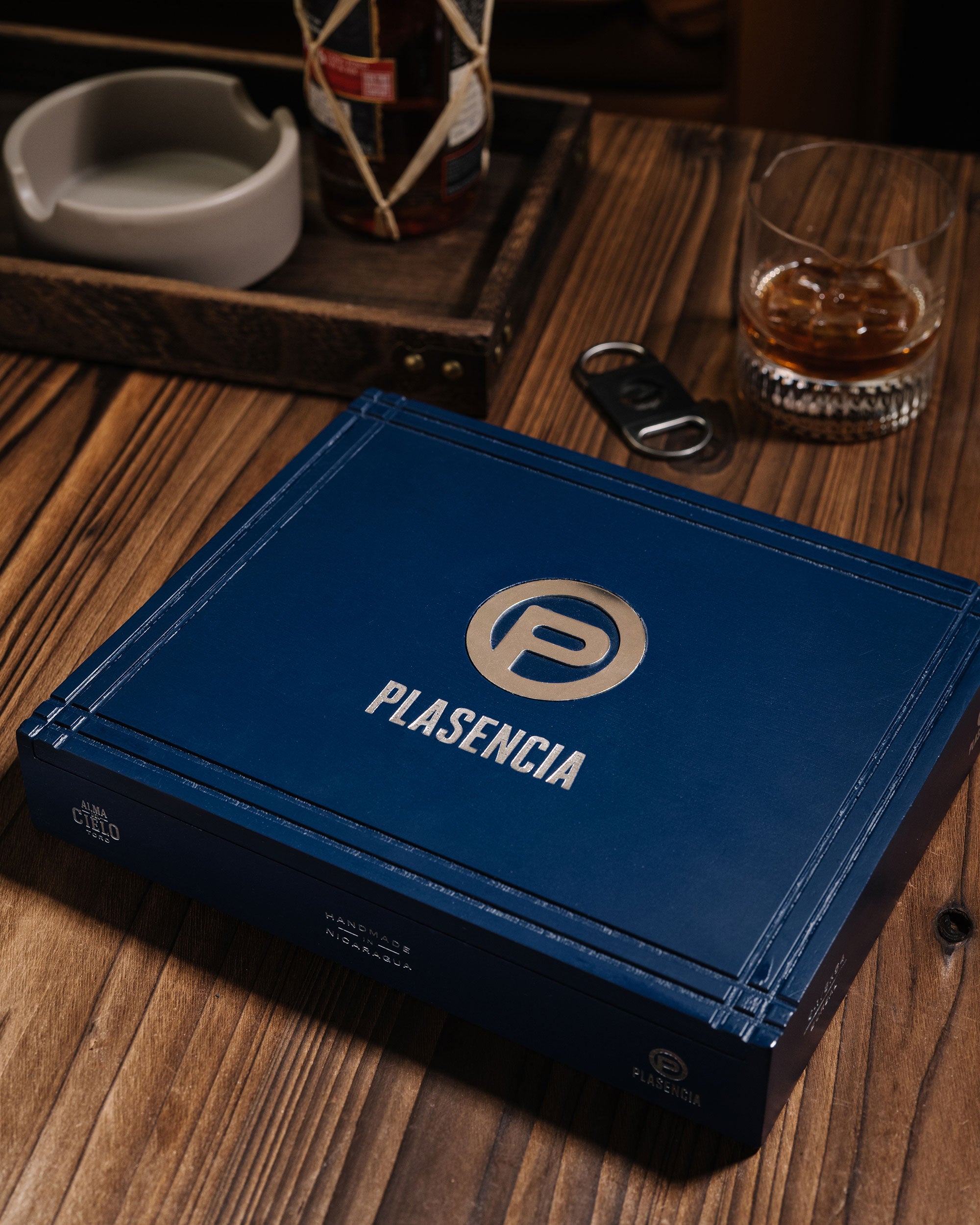 Blue leather-bound book with 'Plasencia' logo on a wooden table with a glass of whiskey and ashtray.