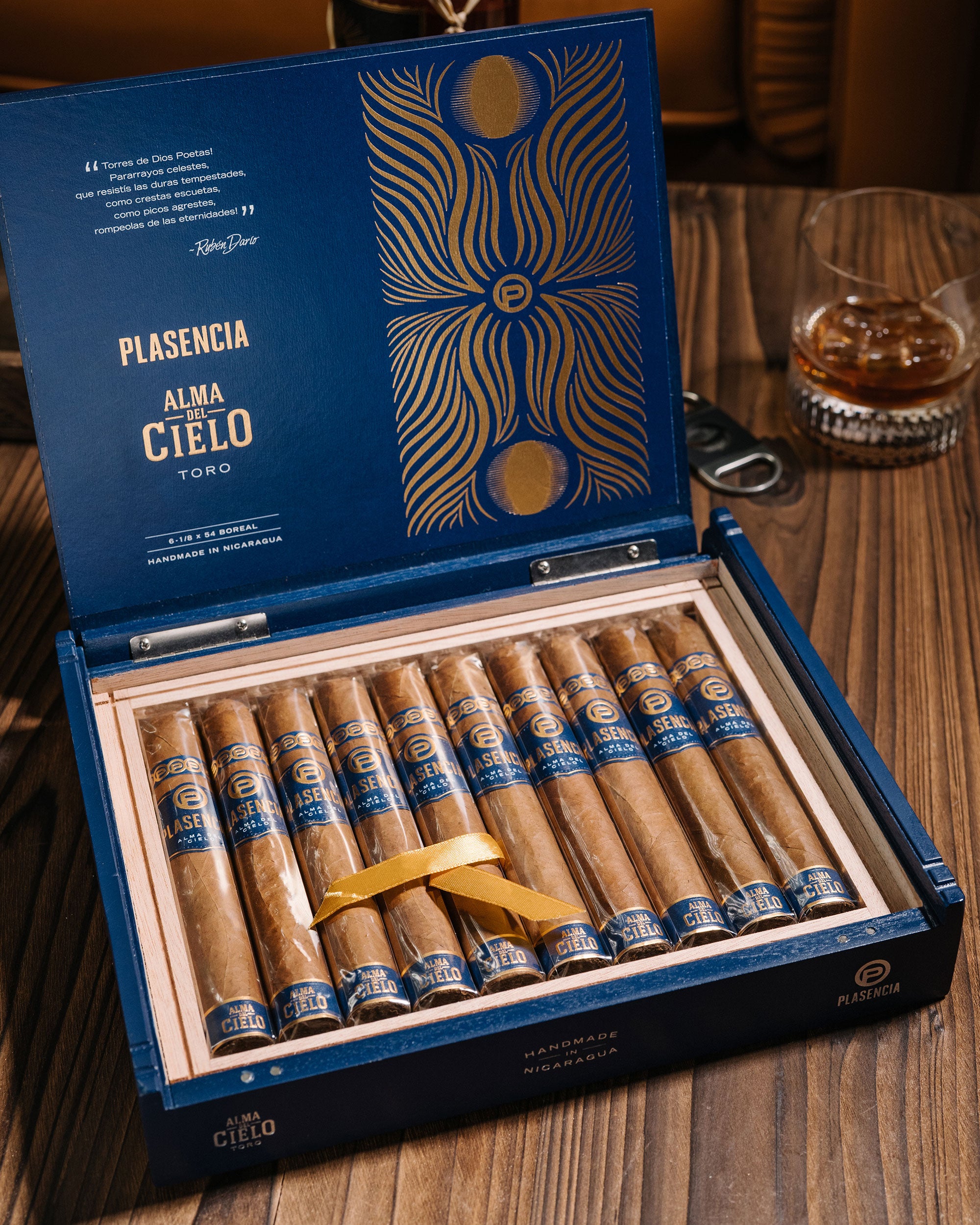 Open box of Plasencia cigars on a wooden table with a glass of whiskey in the background.