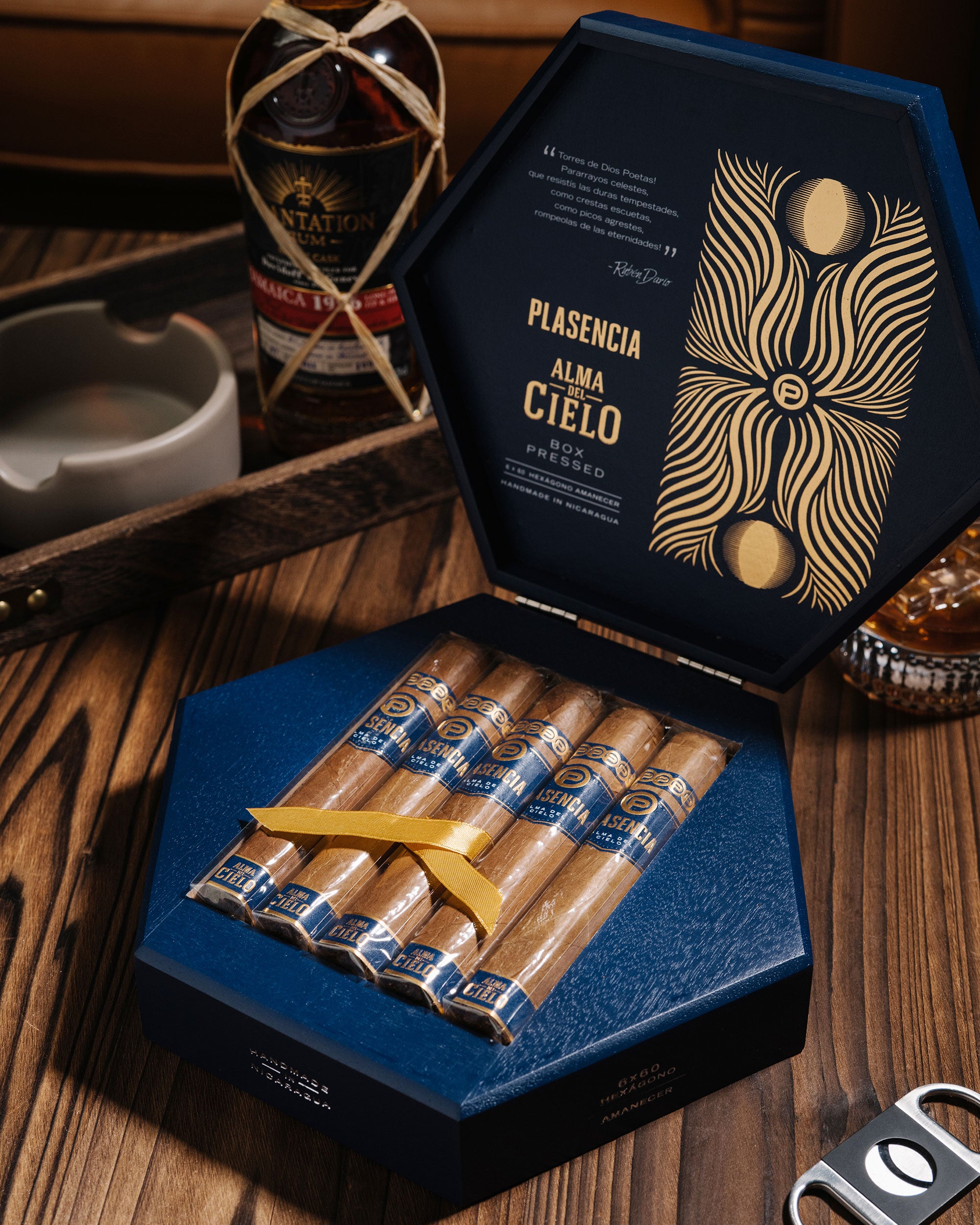 Open box of Plasencia cigars on a wooden table with a bottle of alcohol in the background.