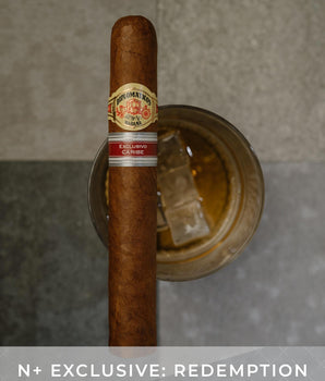Cigar with a label on top of a glass of whiskey, against a neutral background.