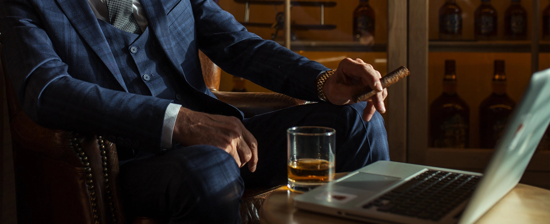 Man in a suit sitting at a bar with a glass of whiskey and a laptop.