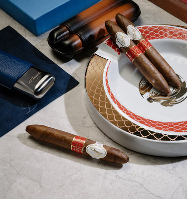 Cigars with decorative bands on a white plate, surrounded by a blue box and a humidor.