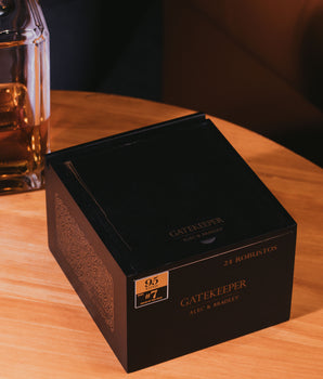 Black 'Gatekeeper' box on a wooden surface with a blurred glass of whiskey in the background.