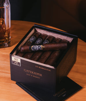 Box of Alec & Bradley Gatekeeper cigars on a wooden table with a glass of beer.