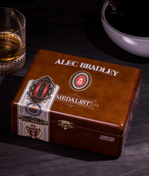 Alec Bradley Medalist cigar box on a dark surface with a glass of whiskey and bowl in the background.