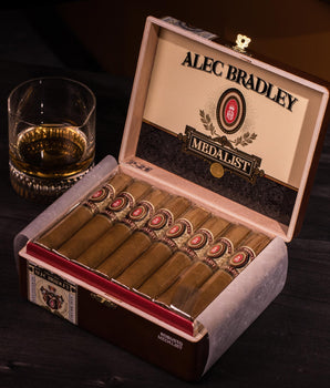 Alec Bradley Medallist cigars in an open humidor with a glass of whiskey on a dark surface.