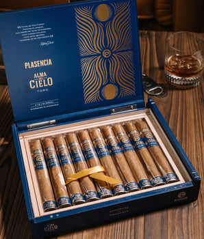 Open box of Plasencia cigars on a wooden table with a glass of whiskey in the background.