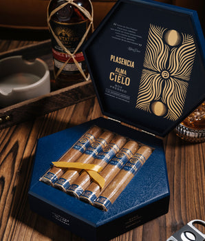 Open box of Plasencia cigars on a wooden table with a bottle of alcohol in the background.