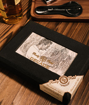 Rocky Patel Decade cigar box on a wooden table with a bottle of whiskey and a spoon in the background.