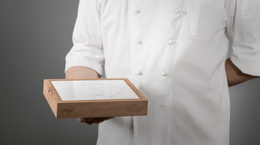 Person in a white coat holding a wooden box with a clear lid against a gray background